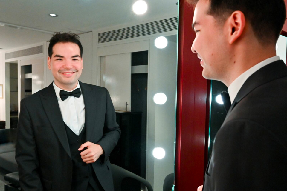 A man wearing a smart suit, looking at a mirror, smiling at the camera.
